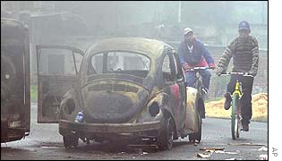 A burnt out car sits by the roadside as activists on bikes cycle past