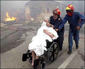 Woman in wheelchair shields her face from smoke as she is wheeled away from fire by firefighters