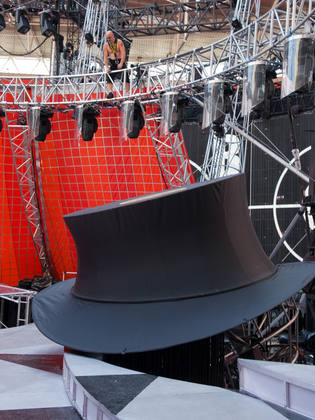 A little man in a harness looks down at a giant top hat.