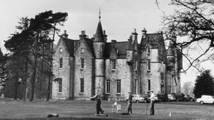 Black and white view of three men playing golf, with a large Scottish Baronial house and trees behind.