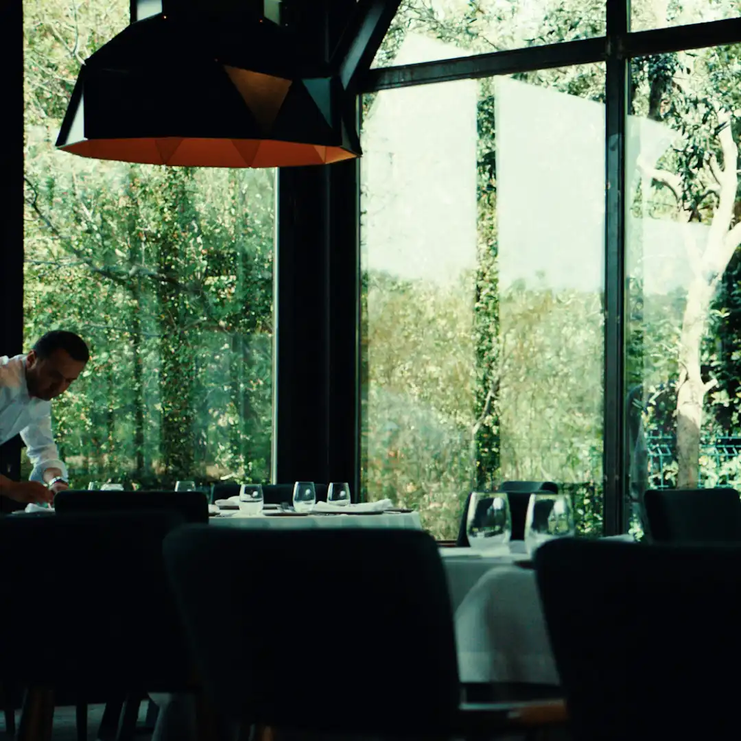 Waiter setting a table inside a modern glass-walled restaurant, surrounded by leafy trees outside.