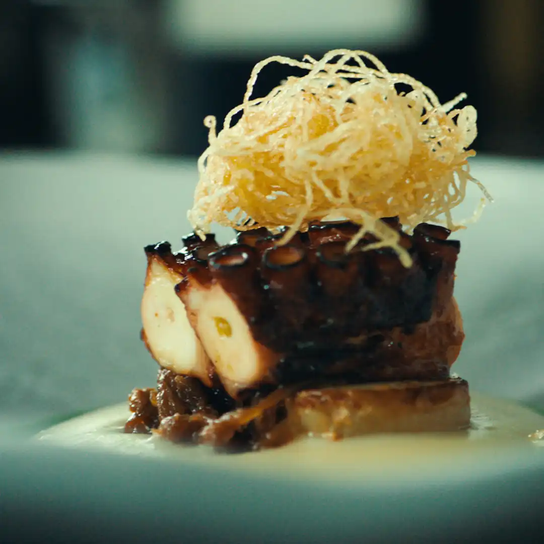 Plated octopus dish topped with crispy golden potato threads, served on a bed of creamy sauce and caramelised onions.