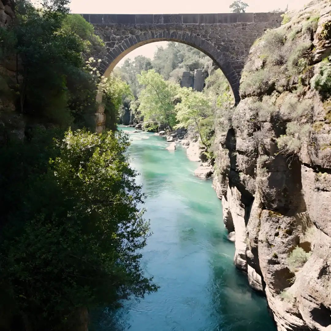 An arched stone bridge spans a turquoise river winding between high canyon walls and green trees.