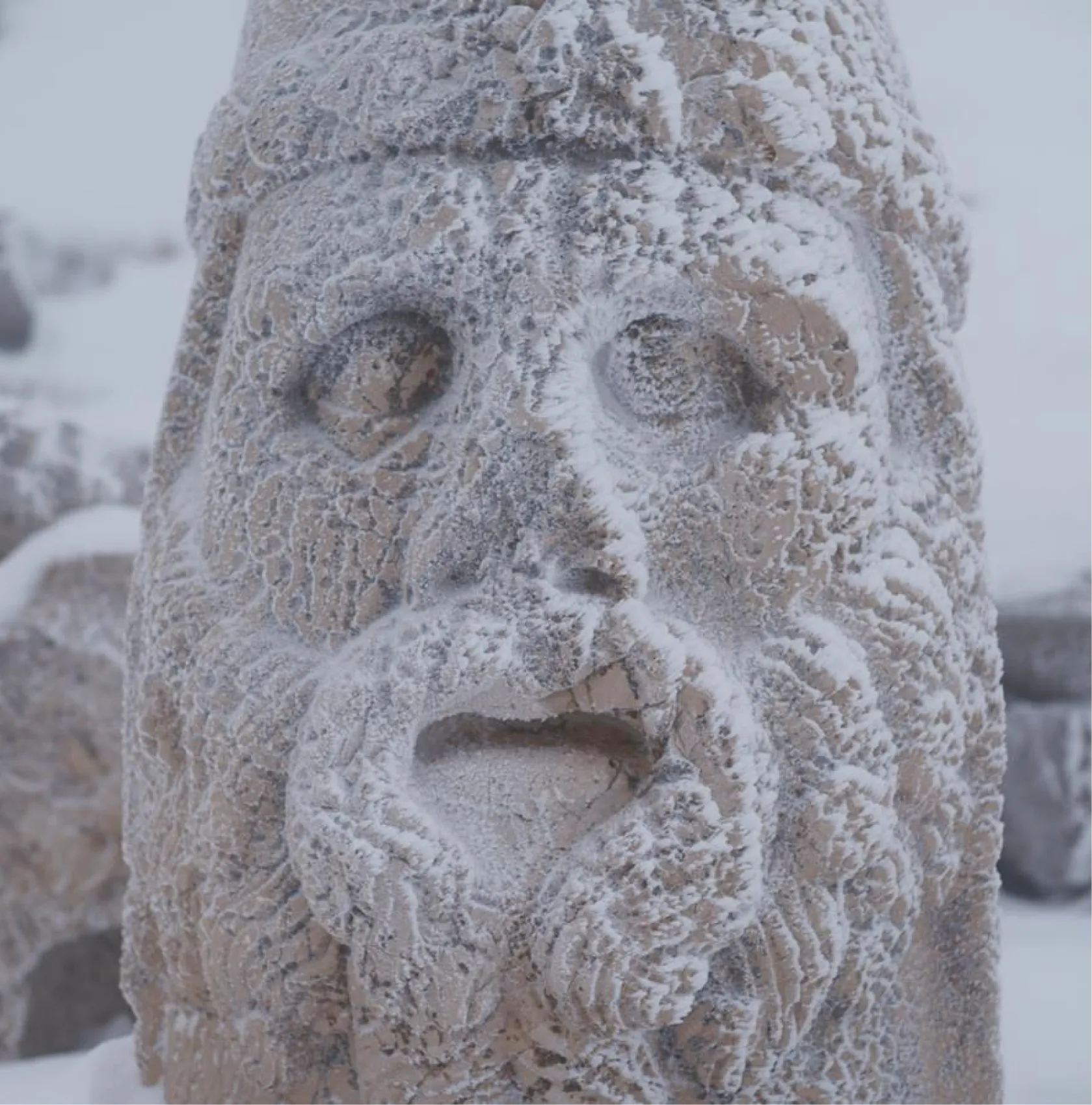 Frost-covered stone head statue at Mount Nemrut, partially buried in snow with intricate headdress detail visible.