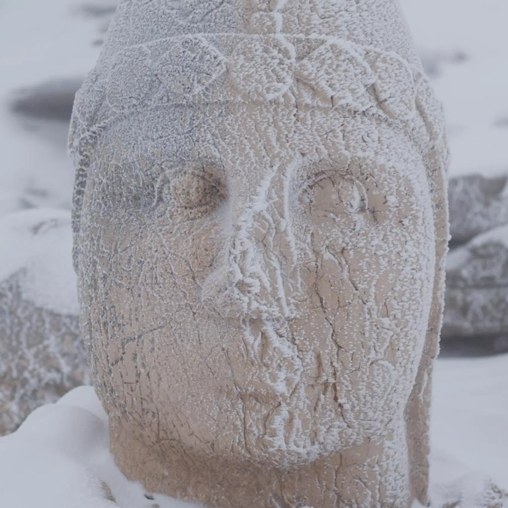 Eroded and snow-covered stone head sculpture with soft facial features, located among the ruins of Mount Nemrut.