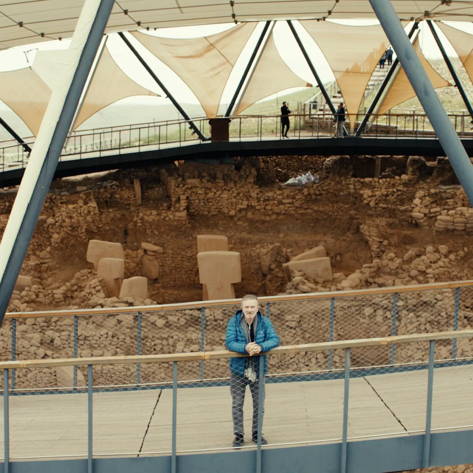 Man standing on a wooden viewing platform overlooking the archaeological excavation at Göbekli Tepe under a protective canopy.